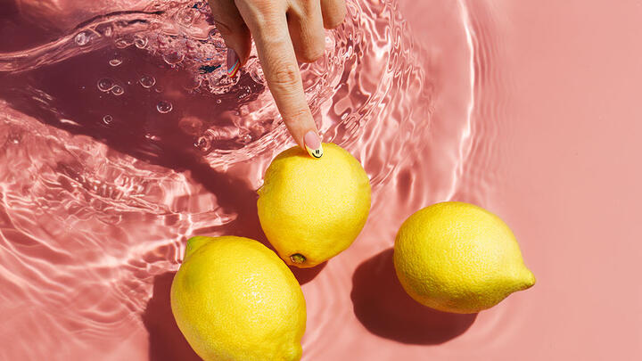 Hand with manicured nails touching one of the three lemons in the water, on a pink surface